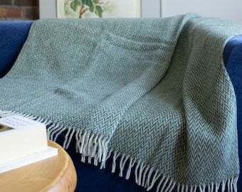 Può includere: Una coperta in lana a spina di pesce verde e bianca con frange su un divano blu. La coperta è piegata sullo schienale del divano e drappeggiata sul bracciolo.