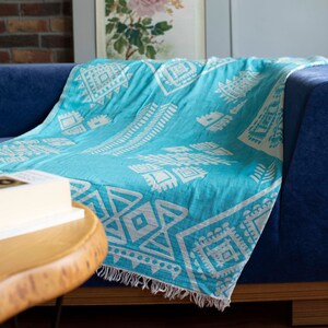 May include: A turquoise blue throw blanket with a white geometric pattern. The blanket is draped over a blue sofa with a wooden frame. A book is on a wooden table in the foreground.