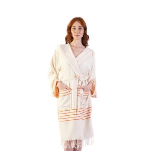 May include: Cream-colored hooded robe with orange horizontal stripes and fringe detail. The robe has a belt, pockets, and is made of a textured fabric. The sleeves have orange accents. A comfortable and stylish spa or beachwear item.