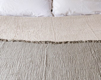 May include: A close-up of a white and beige textured throw blanket with a fringe edge. The blanket is folded in half and the top layer is slightly pulled back to reveal the bottom layer.