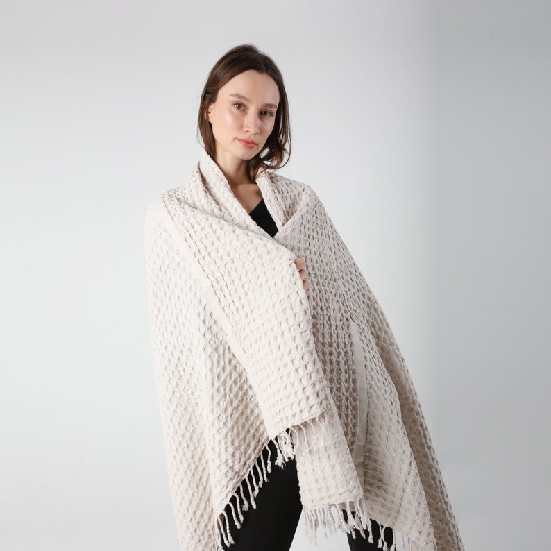 May include: A white waffle weave throw blanket with fringe. The blanket is draped over a person's shoulders.