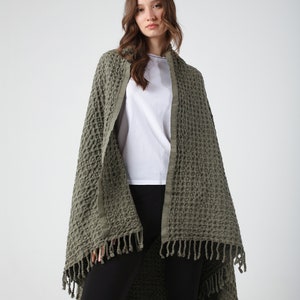 May include: A green waffle weave throw blanket with fringe. The blanket is draped over a person wearing a white t-shirt and black pants.