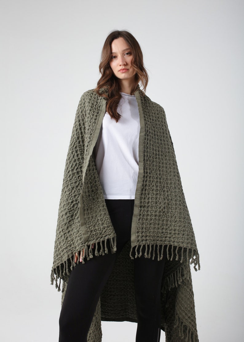 May include: A green waffle weave throw blanket with fringe. The blanket is draped over a person wearing a white t-shirt and black pants.