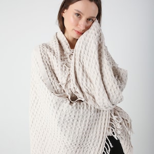 May include: A cream-colored waffle weave throw blanket with fringe. The blanket is draped over a person's shoulders.