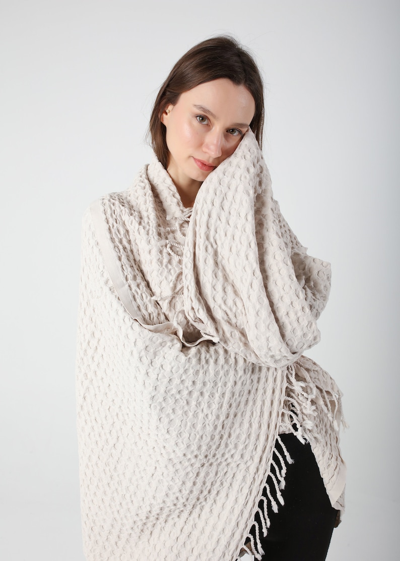 May include: A cream-colored waffle weave throw blanket with fringe. The blanket is draped over a person's shoulders.