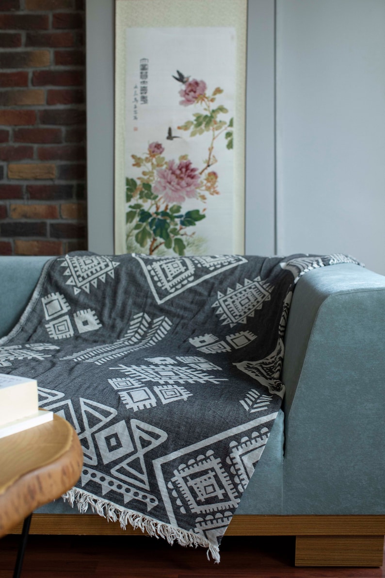 May include: A gray and white patterned throw blanket with fringe edges draped over a light blue sofa. The blanket features a geometric design with white accents on a dark gray background.