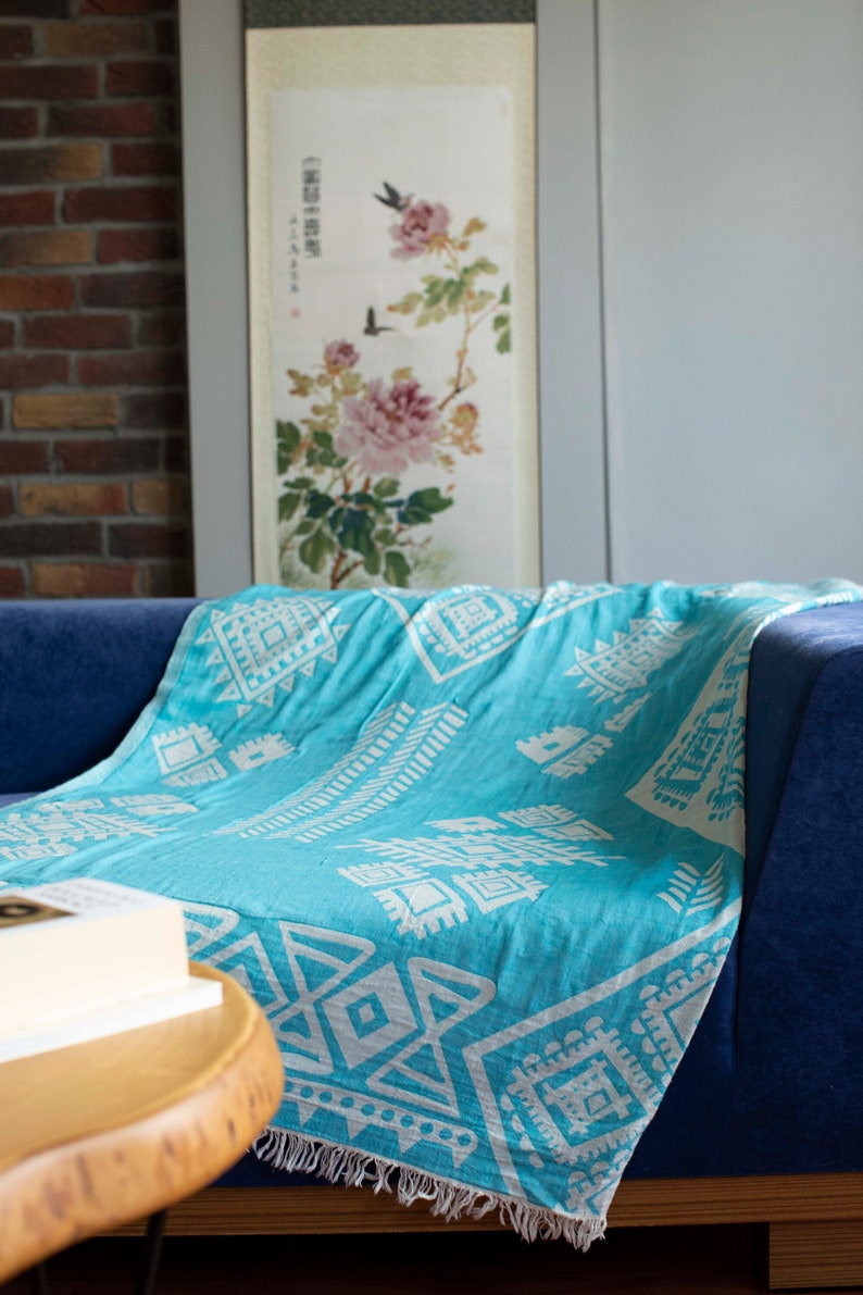 May include: A turquoise blue throw blanket with a white geometric pattern. The blanket is draped over a blue sofa with a wooden frame. A book is on a wooden table in the foreground.