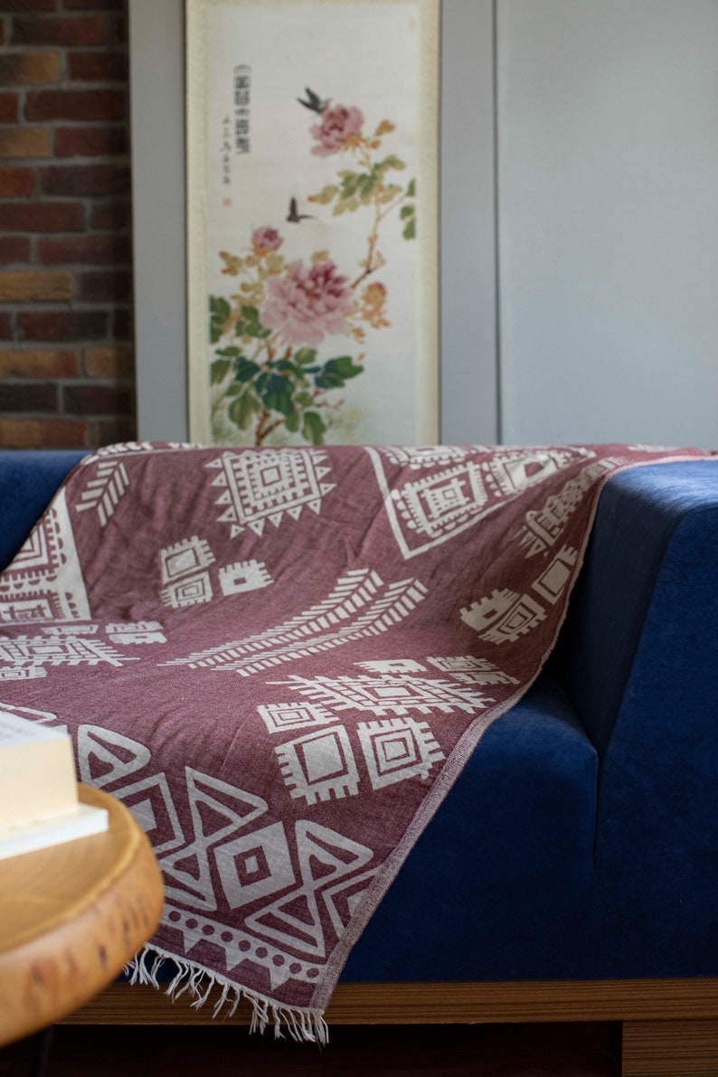 May include: A burgundy throw blanket with a white geometric pattern is draped over a blue sofa. The blanket has a fringe border.