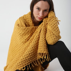 May include: A person wearing a yellow waffle weave throw with fringe. The throw is draped over the person's shoulders and they are sitting on the floor.