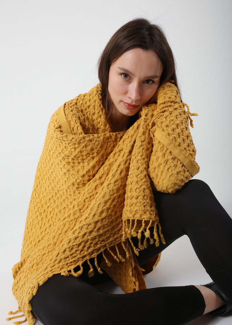 May include: A person wearing a yellow waffle weave throw with fringe. The throw is draped over the person's shoulders and they are sitting on the floor.