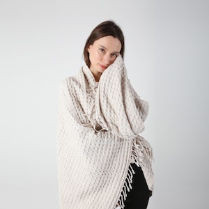 May include: A woman wearing a cream-colored waffle-knit throw with fringe. The throw is draped around her shoulders and she is holding it with one hand.