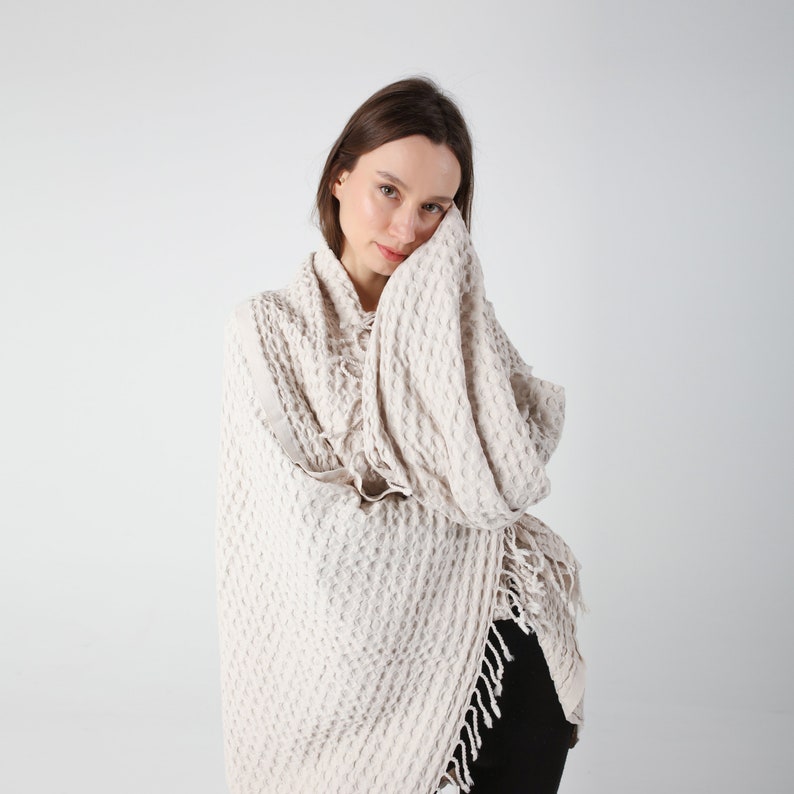 May include: A woman wearing a cream-colored waffle-knit throw with fringe. The throw is draped around her shoulders and she is holding it with one hand.