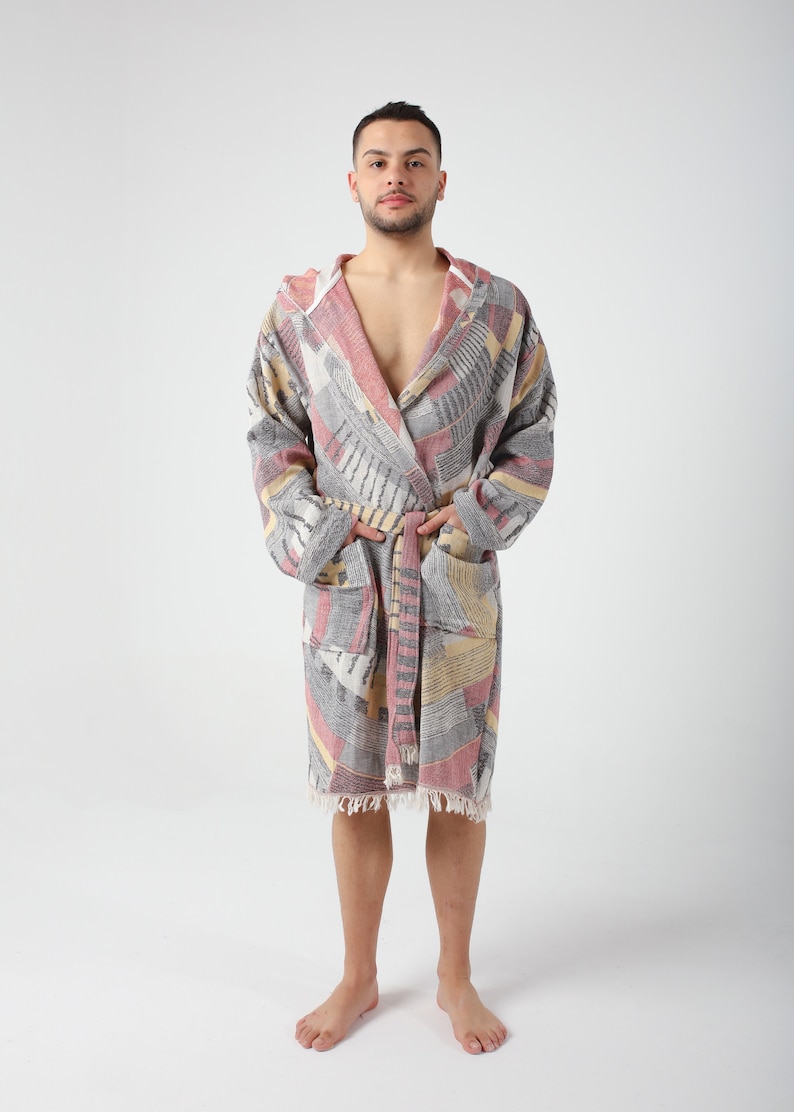 May include: A hooded bathrobe with a geometric pattern in shades of gray, pink, and yellow. The robe has a sash tie and fringe trim.