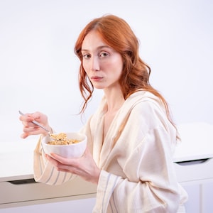 May include: A person wearing a cream-colored robe, holding a white bowl of oatmeal and a spoon. The person has red hair and is seated at a white desk. Small vases with yellow and pink flowers are on the desk.