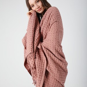 May include: A soft, pink waffle weave throw blanket with fringe. The blanket is draped over a person's shoulders.