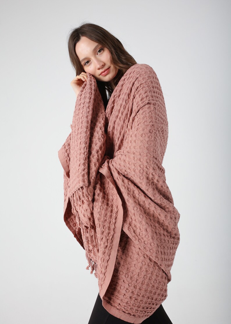 May include: A soft, pink waffle weave throw blanket with fringe. The blanket is draped over a person's shoulders.