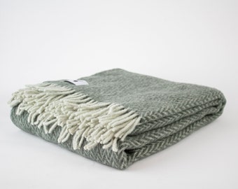 Può includere: Una coperta di lana verde a spina di pesce piegata con frange bianche.