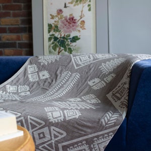 May include: A gray throw blanket with a white geometric pattern is draped over a blue couch. The blanket has a fringe border.