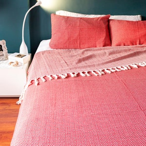 Op de afbeelding: Rode en witte gepatroneerde deken met franjes op een bed. De deken is gemaakt van een zachte, geweven stof.