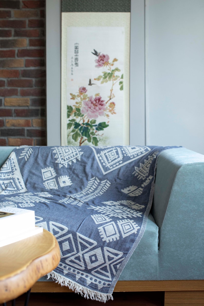 May include: A blue and white patterned throw blanket with a fringe edge draped over a light blue sofa. The blanket has a geometric design with white triangles and squares on a blue background. The sofa is in front of a wall with a framed painting of flowers and birds.