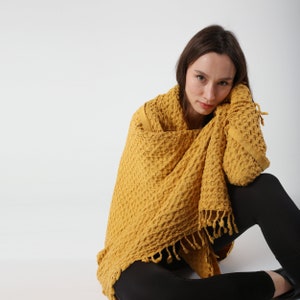 May include: A woman sits with a yellow waffle weave throw draped around her shoulders. The throw has a fringe border.