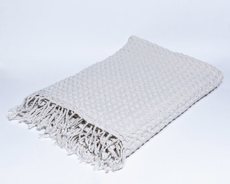 May include: A white waffle weave throw blanket with a fringe border. The blanket is folded in half and lying on a white surface.