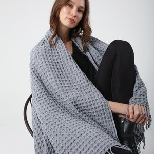 May include: A gray waffle weave throw blanket with fringe. The blanket is draped over a person sitting on a chair.