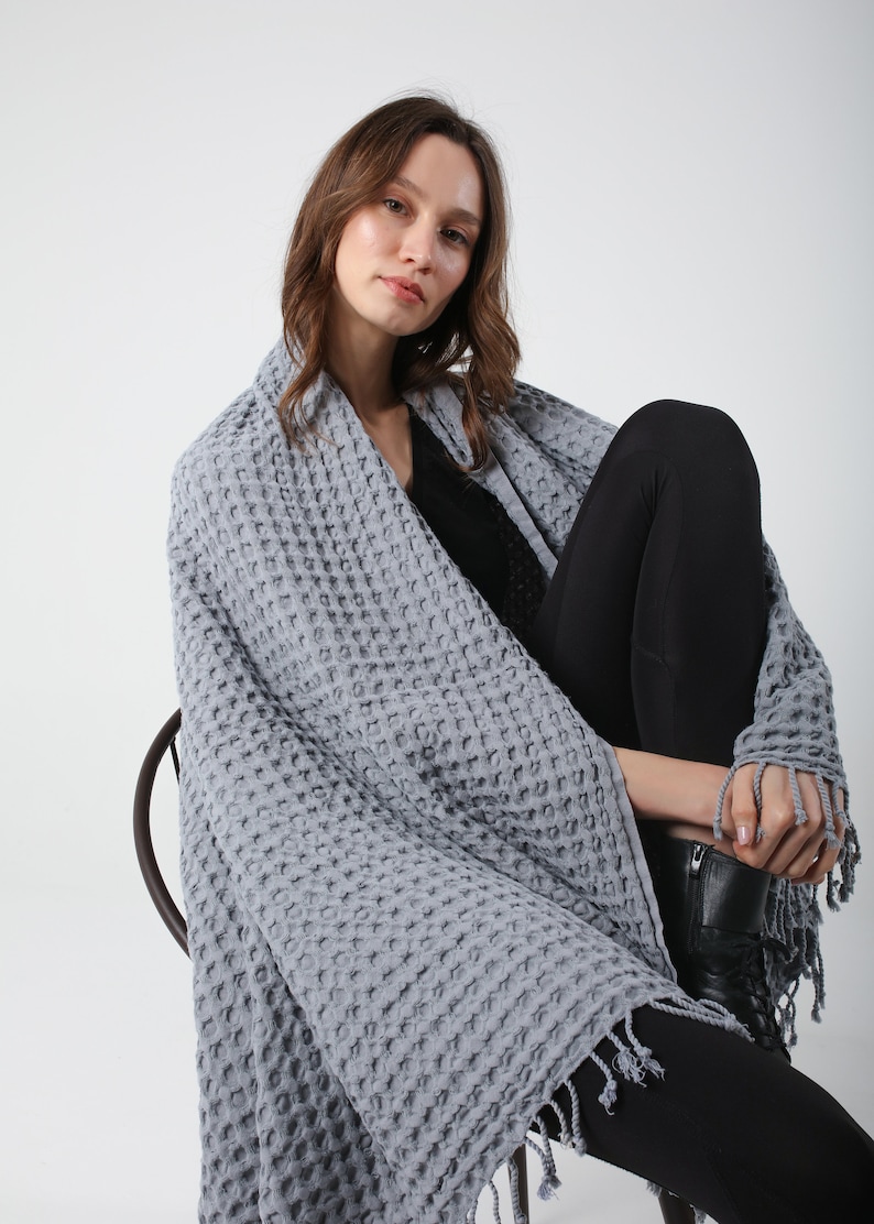 May include: A gray waffle weave throw blanket with fringe. The blanket is draped over a person sitting on a chair.