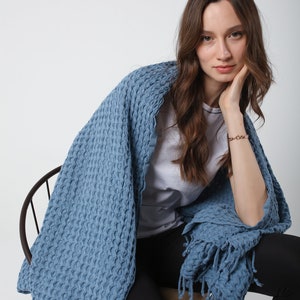 May include: A woman sitting on a chair wearing black pants and a white t-shirt, wrapped in a blue waffle-textured throw blanket with fringe.