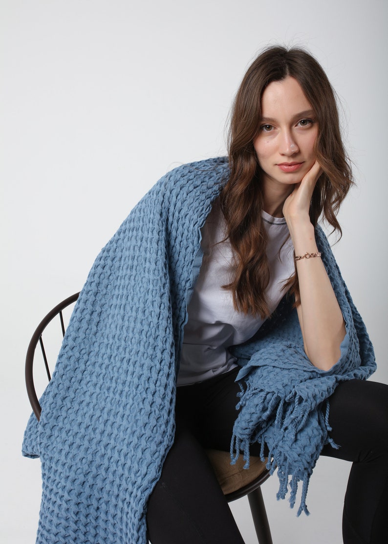 May include: A woman sitting on a chair wearing black pants and a white t-shirt, wrapped in a blue waffle-textured throw blanket with fringe.