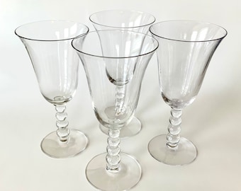 May include: Four clear glass wine glasses with beaded stems. The glasses are arranged in a semi-circle on a white surface.