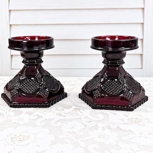 May include: A pair of deep red glass candle holders featuring a wide, circular top rim, a short neck, and a hexagonal base. The glass surface is decorated with a raised, textured diamond pattern and circular accents throughout the exterior.