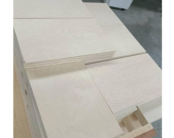 May include: A stack of light wood panels, likely plywood, with a smooth, light-colored surface. The panels are stacked neatly, showing the grain of the wood.