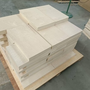 May include: A stack of light wood panels, probably plywood, are stacked on a wooden pallet. The panels are smooth and have a light wood grain.