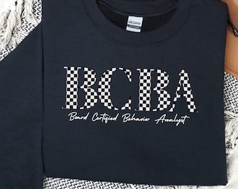 May include: A black crewneck sweatshirt with the letters "BCBA" in a checkered pattern. Below the letters, the words "Board Certified Behavior Analyst" are written in a script font. The sweatshirt is displayed on a white and black patterned blanket.