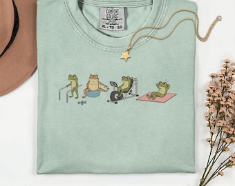 May include: A light green Comfort Colors t-shirt with a crew neck and short sleeves. The shirt features a graphic of frogs exercising, including weightlifting, using a balance ball, riding a stationary bike, and relaxing on a yoga mat. A gold star pendant necklace is draped across the shirt.