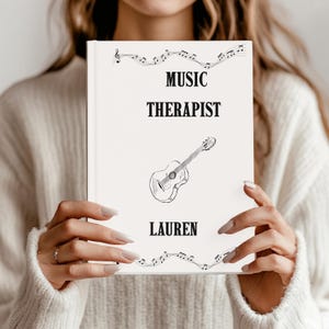 Peut inclure: Un livre blanc avec les mots "MUSIC THERAPIST" en gras noir, et le nom "LAUREN" en dessous. Un croquis d'une guitare est au centre. Des notes de musique et une clé de sol sont en haut. Le livre est tenu par une personne.