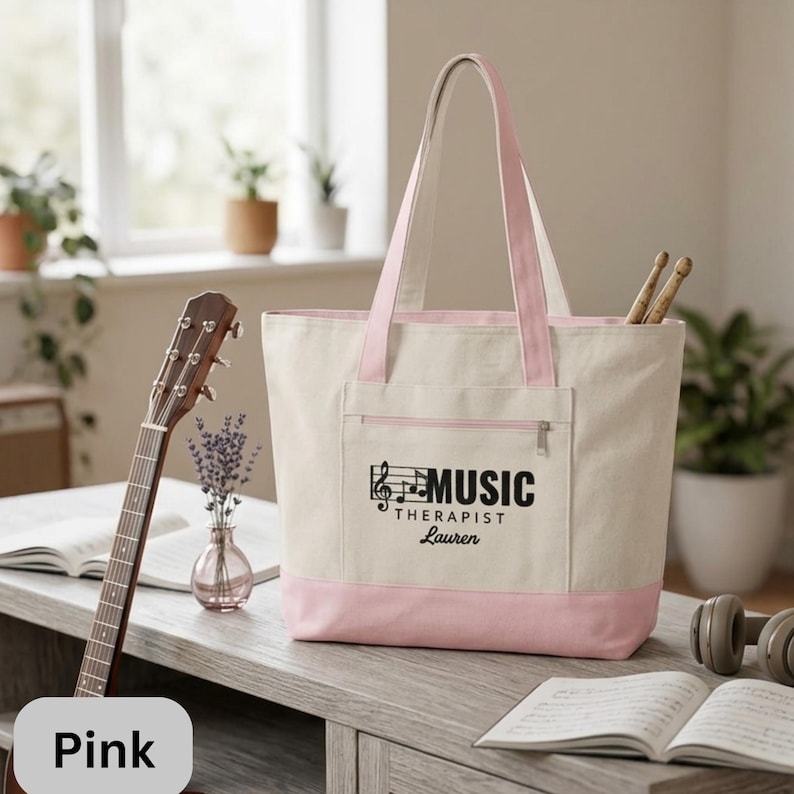 May include: A canvas tote bag with a pink base and straps. The bag has a front pocket with a zipper and the words "MUSIC THERAPIST Lauren" printed in black. A guitar and drumsticks are visible.