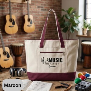 May include: Cream-colored tote bag with maroon accents, featuring a front pocket and handles. The bag displays the text "MUSIC THERAPIST Lauren" with a musical note graphic. Guitars and drums are visible in the background.