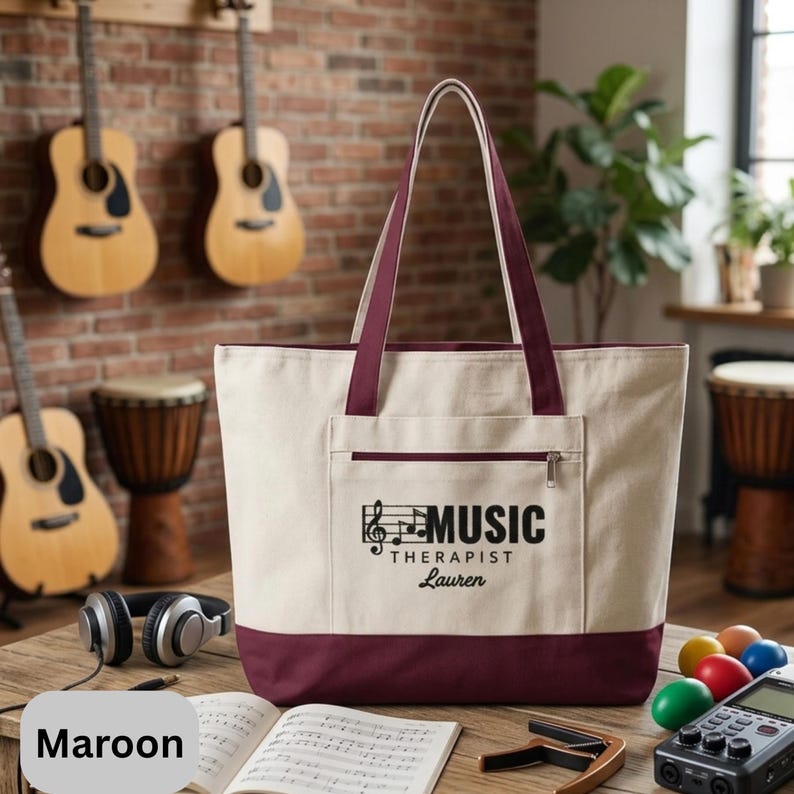 May include: Cream-colored tote bag with maroon accents, featuring a front pocket and handles. The bag displays the text "MUSIC THERAPIST Lauren" with a musical note graphic. Guitars and drums are visible in the background.