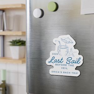 May include: A white magnet with a blue illustration of a tropical drink, starfish, and umbrella. Text reads "2026 Last Sail Before The Veil Erica's Bach Trip". The magnet is on a silver refrigerator door.