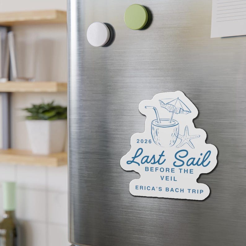 May include: A white magnet with a blue illustration of a tropical drink, starfish, and umbrella. Text reads "2026 Last Sail Before The Veil Erica's Bach Trip". The magnet is on a silver refrigerator door.