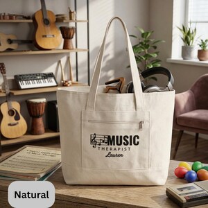 May include: A natural canvas tote bag with a front pocket and zipper. The bag features the text "MUSIC THERAPIST Lauren" in black, along with a musical note design. The bag is filled with headphones and other items.