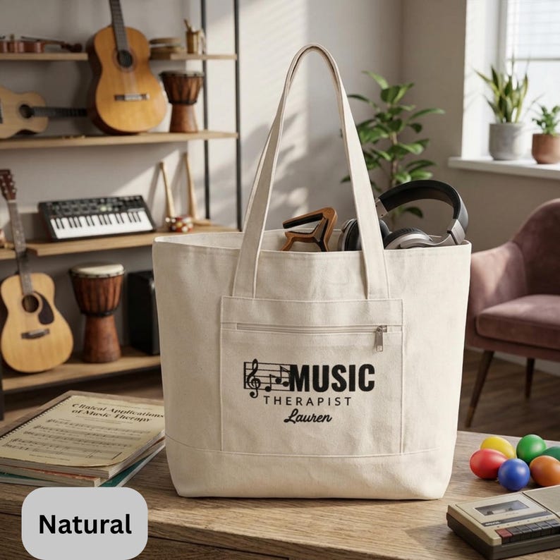 May include: A natural canvas tote bag with a front pocket and zipper. The bag features the text "MUSIC THERAPIST Lauren" in black, along with a musical note design. The bag is filled with headphones and other items.