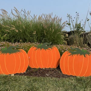 Puede incluir: Tres decoraciones de calabazas de madera naranja con tallos verdes, exhibidas en un césped. Las calabazas tienen ranuras verticales y están colocadas sobre un fondo de vegetación y un cielo azul claro.