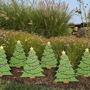 Könnte beinhalten: Sechs grüne Weihnachtsbaum-Gartendekorationen. Jeder Baum hat einen goldenen Stern und bunte Lichter. Die Bäume stehen in einem Garten mit Blumen und hohem Gras. Die Bäume sind etwa 60 cm hoch.