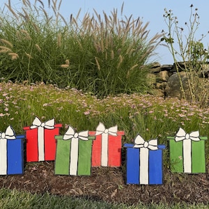Puede incluir: Seis decoraciones de cajas de regalo de madera de colores con lazos blancos y rayas verticales. Las cajas están pintadas en rojo, verde y azul, y están colocadas en un jardín con flores y hierba alta.