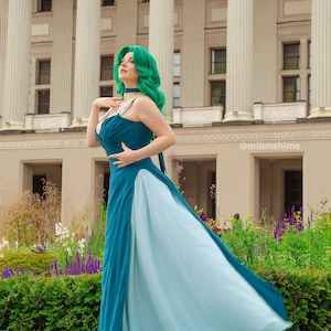 Puede incluir: Un vestido de noche azul verdoso y azul claro con tirantes finos y una falda fluida. La modelo lleva una peluca verde y una gargantilla. El fondo presenta un edificio con columnas y un jard&iacute;n.