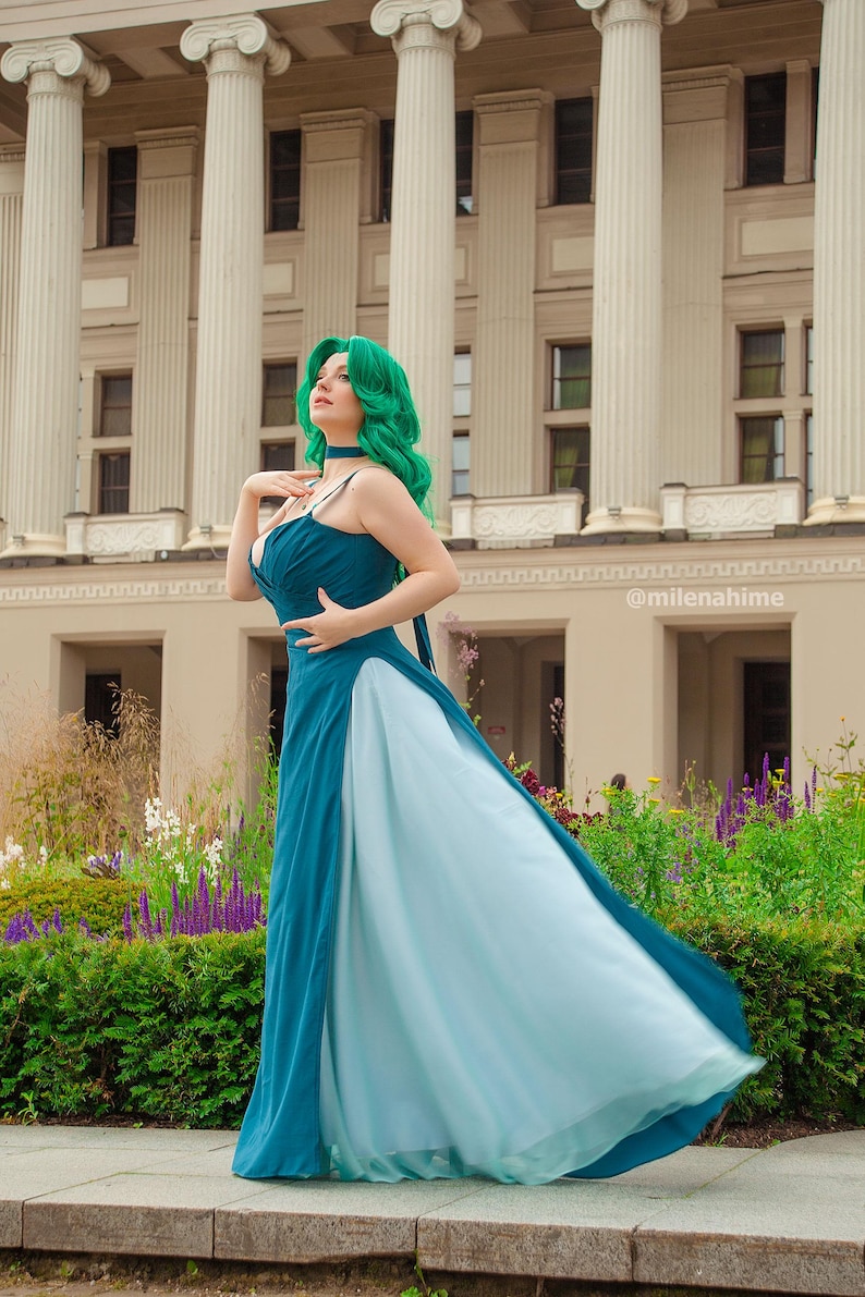 Puede incluir: Un vestido de noche azul verdoso y azul claro con tirantes finos y una falda fluida. La modelo lleva una peluca verde y una gargantilla. El fondo presenta un edificio con columnas y un jard&iacute;n.