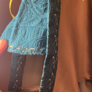 May include: A teal blue fabric with a gold trim and a gold cord with a gold bead at the end. The fabric has a textured pattern.